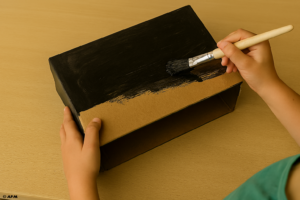 Des mains d'enfant peignent l'extérieur d'une boîte en carton avec de la peinture noire.